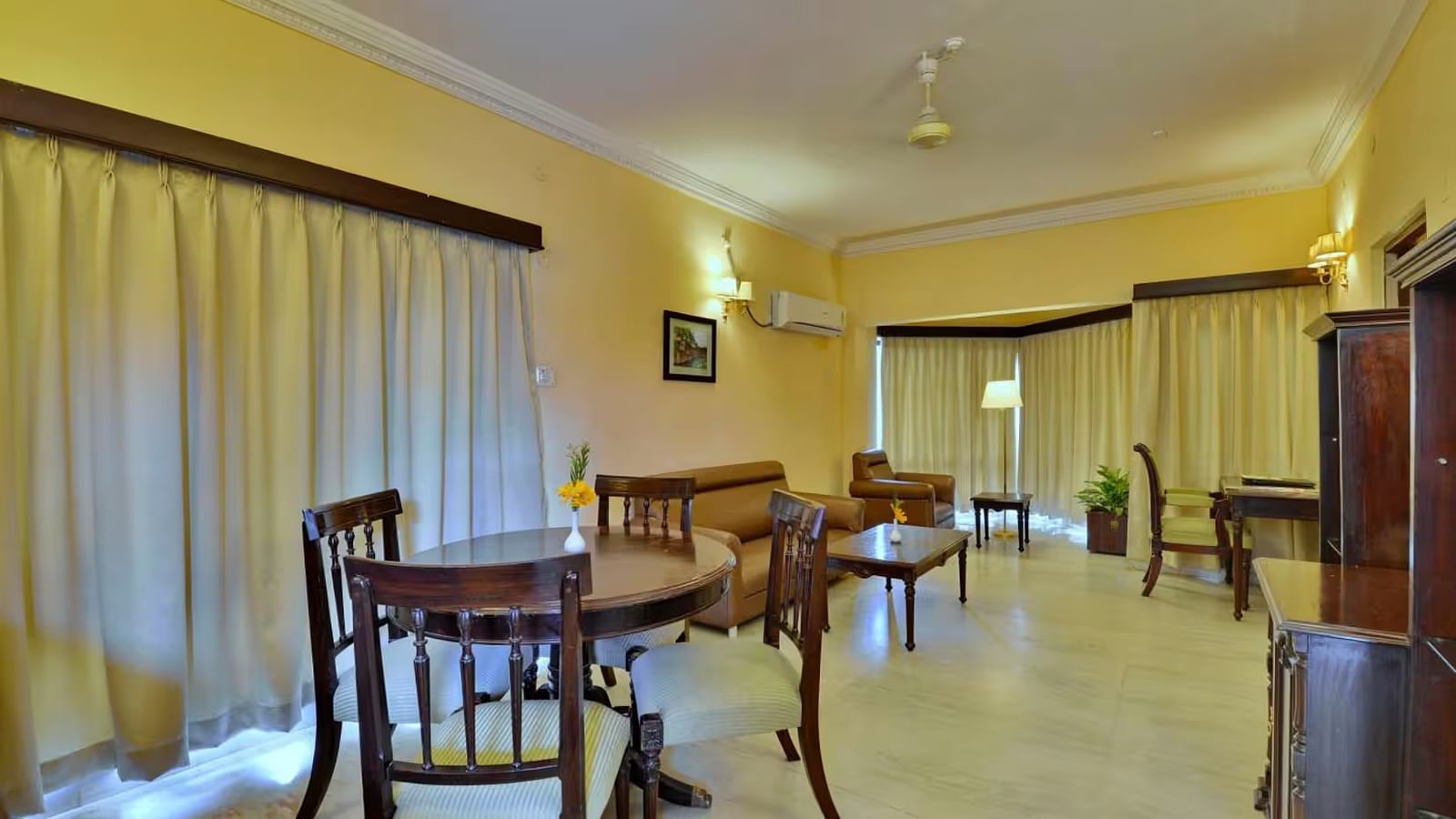  The living area of the suite features a wall-mounted television on a wooden shelf, with furniture arranged opposite it, and two adjacent windows to the side, fully covered by curtains.