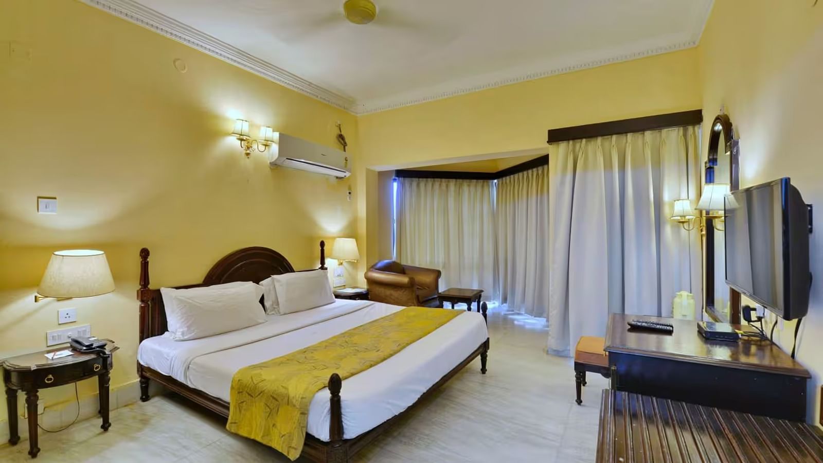  A spacious room with a large bed, a wall mounted television opposite, a dressing table with a chair and wall-mounted mirror beside it, and an air conditioner positioned directly above the bed.