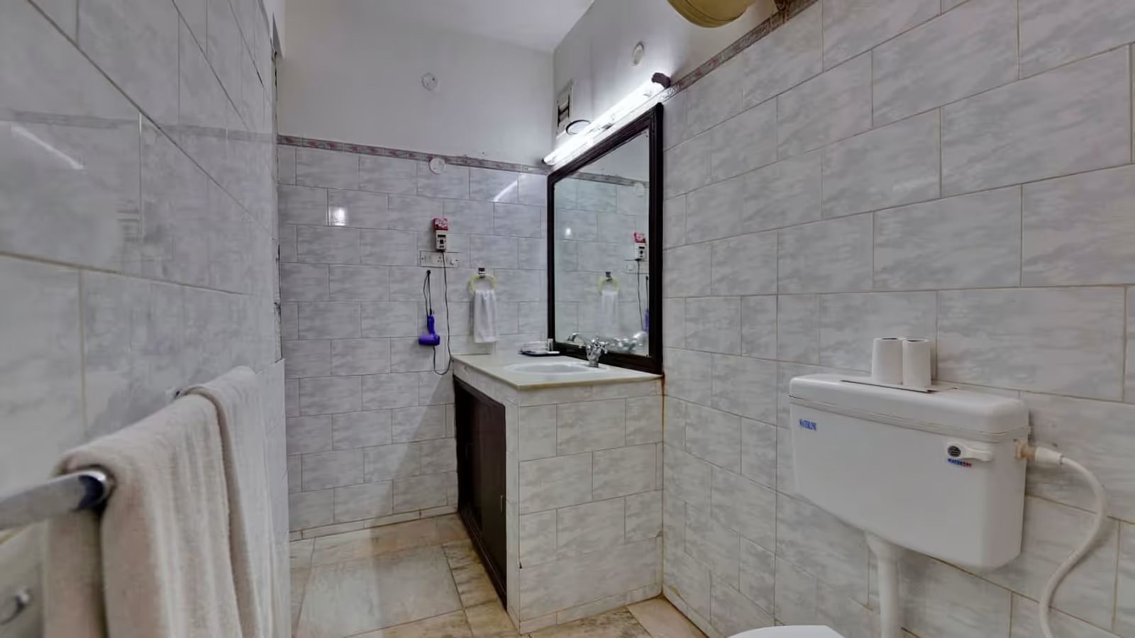 A bathroom with a western toilet, towels hung on a rack, a washbasin with toiletries on the side, and a mirror mounted directly above it.