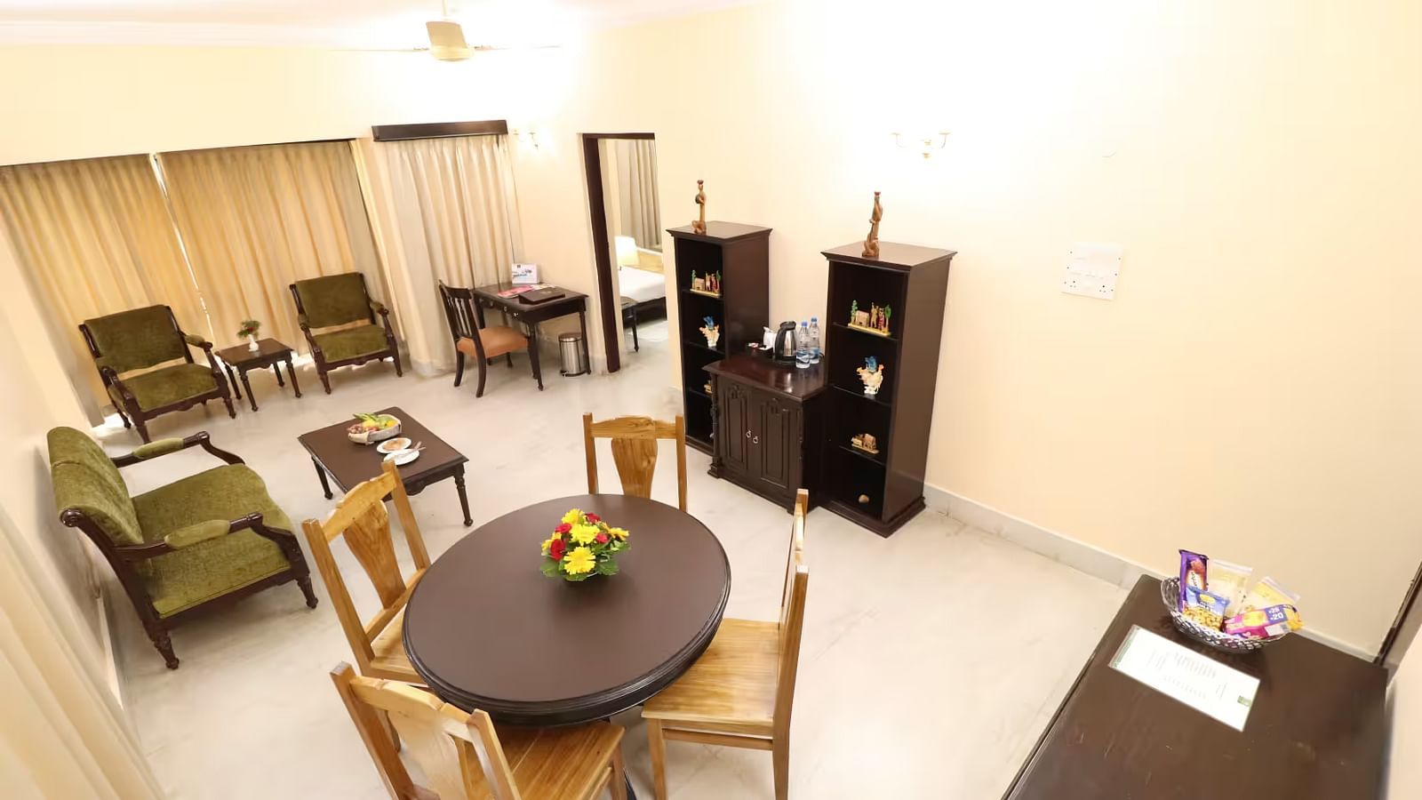  A top view of the suite's separate living area shows a wooden cupboard with decorative items on the side, a circular table with chairs around it, and windows covered by curtains, with additional seating furniture nearby.