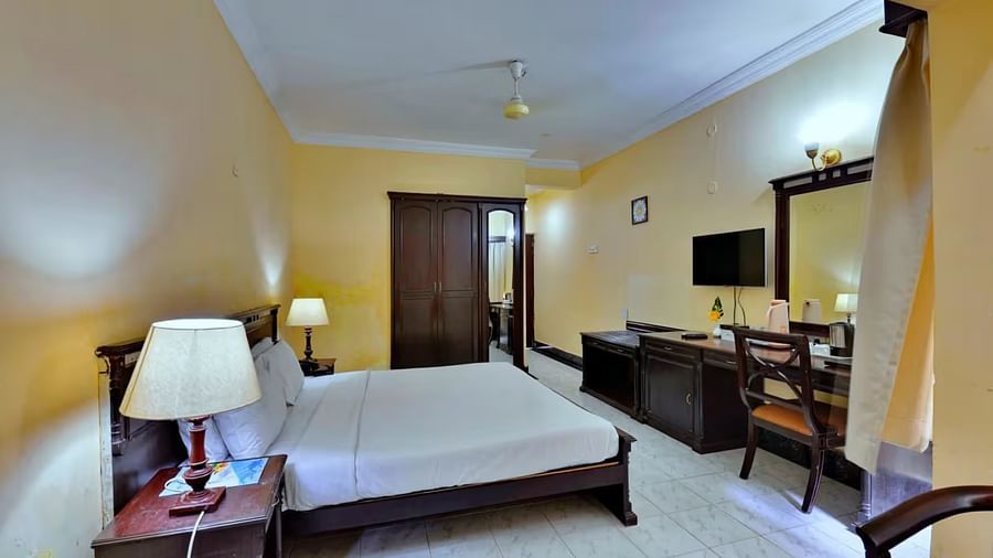  A spacious room with a bed and bedside table with lamps, a wall-mounted shelf adorned with decorative elements, and a sofa, table, and desk positioned on the opposite side.