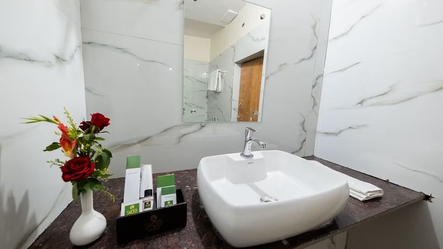  The bathroom of the Standard Room features an elegant washbasin with a mirror above it, a tray beside it with essential toiletries, and a flower vase with an assortment of flowers for decoration.