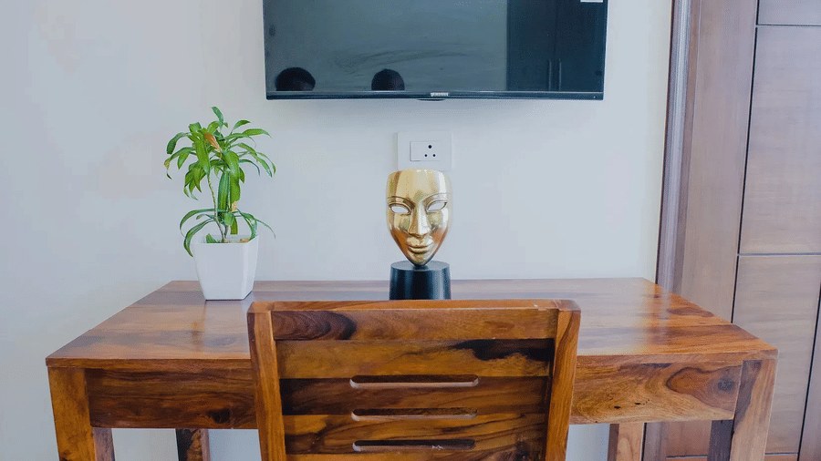 Wooden desk and chair below a wall-mounted TV, with a gold mask ornament and a potted plant