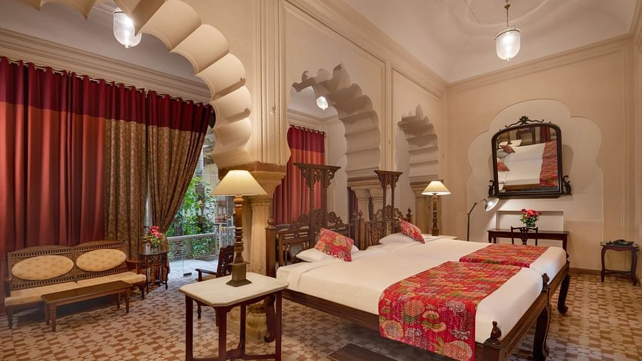 A room with a bed, a sofa, tables with lamps, a mirror, curtains, and arched architectural details at Tijara Fort-Palace - 19th Century, Alwar.