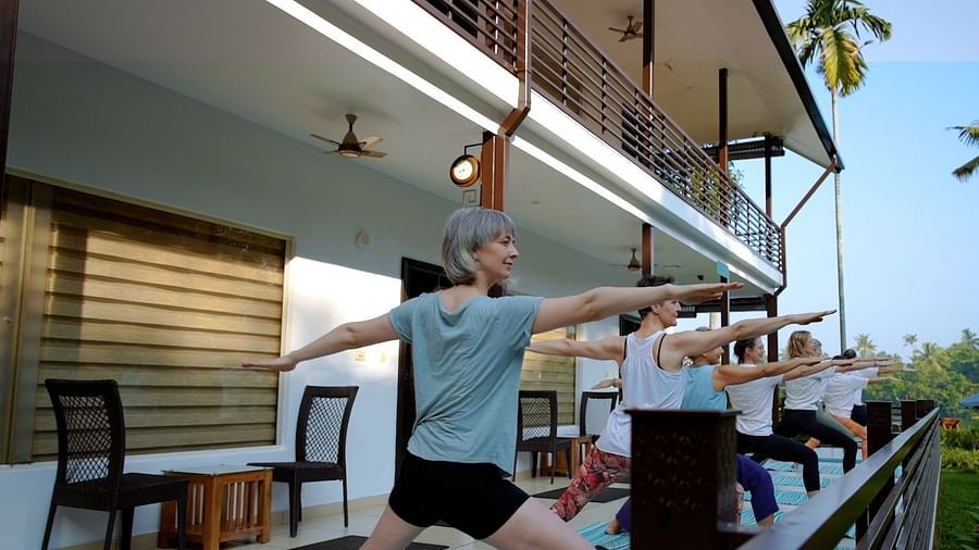 A group of people practice yoga warrior pose on a patio with a building and palm trees in the background.