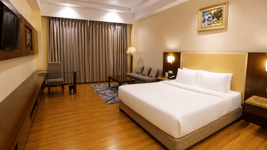 Deluxe room at Golden Tulip Kukas Jaipur featuring a king-sized bed with crisp white bedding, polished hardwood flooring, dark drapes, and warm ambient lighting.