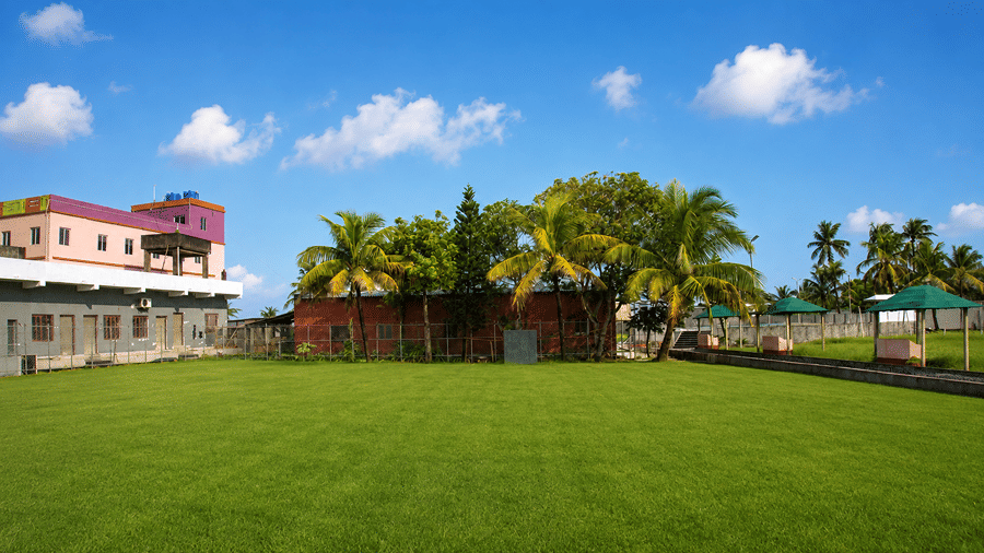 Ground-level view of the lawn at Pride Elite Mandarmani with the hotel side wing, coconut palms, and green canopy seats under a clear blue sky.