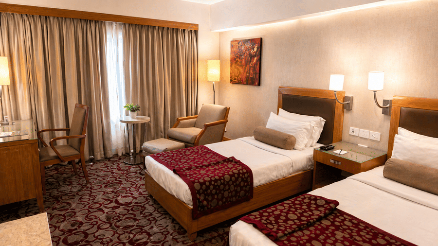 Twin beds with patterned carpet, lamps, and cozy seating area at Pride Plaza Ahmedabad.