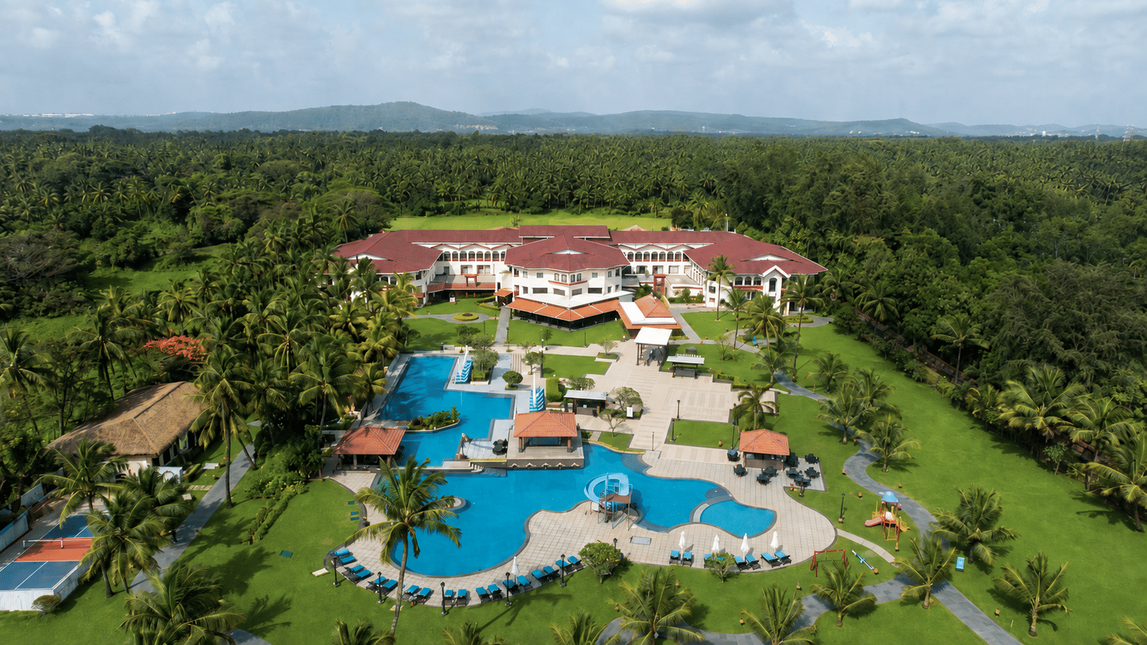 Aerial view of the swimming pools and lush greenery surrounding Kenilworth Resort & Spa, Goa on a sunny day.