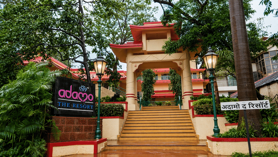 The entrance of the hotel with its red and yellow gate, yellow stairs, and the hotel logo on a signboard on the side while being surrounded by all the trees of the property.