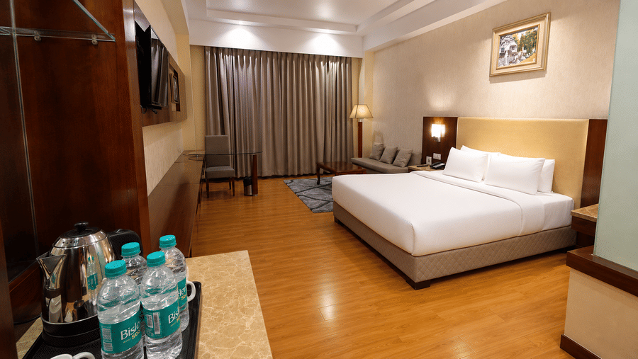 Deluxe room at Golden Tulip Kukas Jaipur with a neatly made king bed, hardwood floors, framed wall art, and complimentary water bottles placed near the bed.