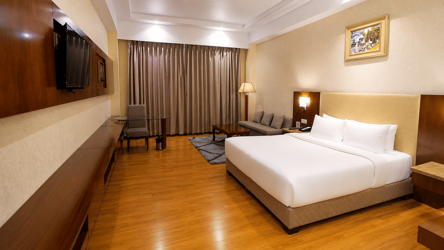 Corner view of the Deluxe room at Golden Tulip Kukas Jaipur featuring a king bed, two armchairs along the wall, dark drapes, and warm ceiling lighting.