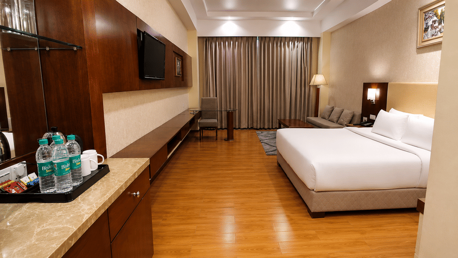 Deluxe room at Golden Tulip Kukas Jaipur with a king-sized bed, wooden desk stocked with water bottles, dark floor-length curtains, and warm overhead lighting.