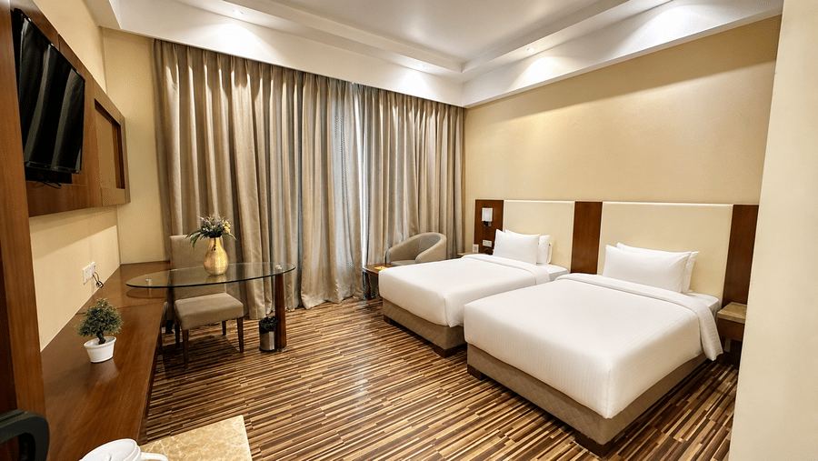 Superior Twin room at Golden Tulip Kukas Jaipur featuring two single beds, wall-mounted TV, grey curtains, striped carpet, and warm recessed ceiling lighting.