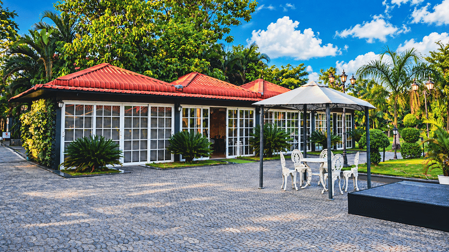 Red-roofed glass-walled cottages beside a paved courtyard with a white gazebo, garden chairs and palm trees at HM Hotel & Resort, Dibrugarh.