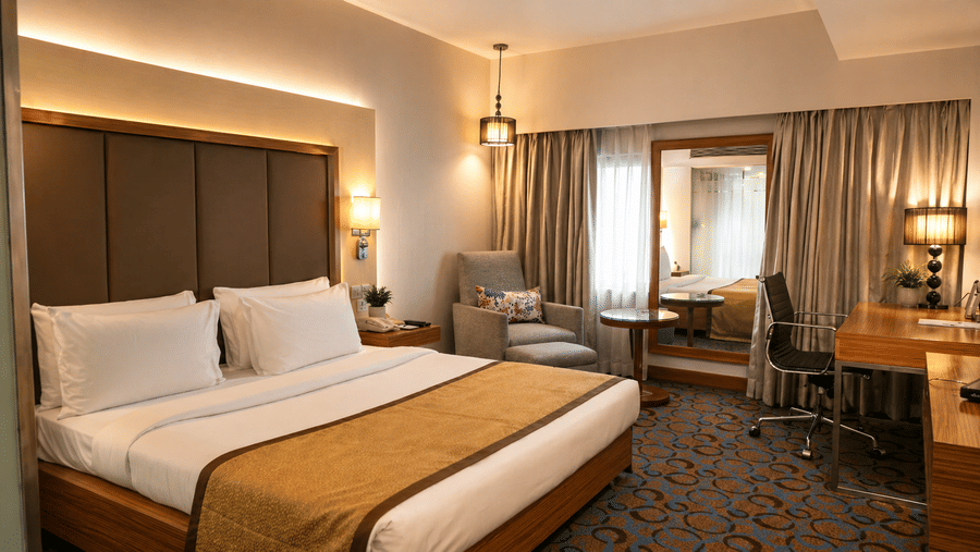 King bed with crisp linens, bedside lamps, desk, and window with curtains at Pride Plaza Ahmedabad.