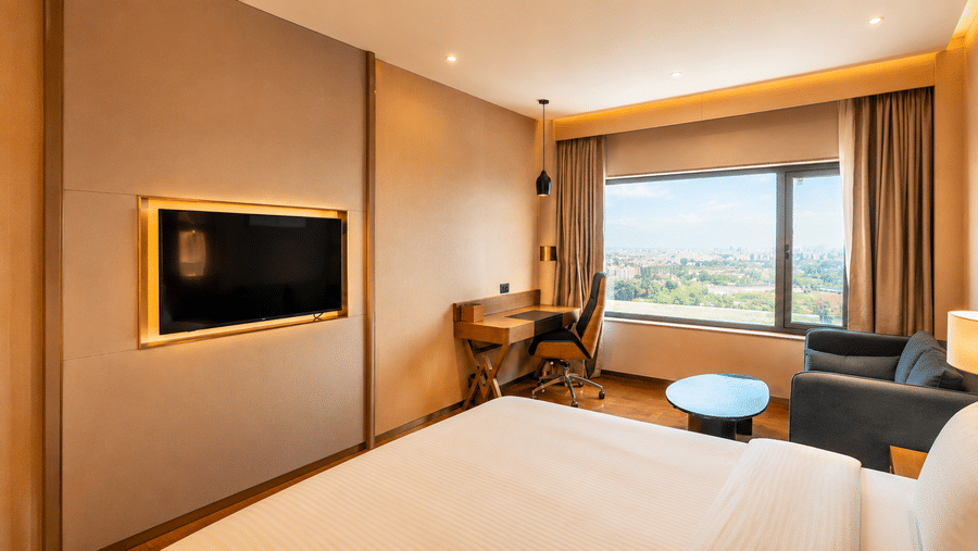 Superior King Room at Pride Plaza Indore showing a bed, a wall-mounted panel, a writing desk with a chair, a sofa, and a large window view.
