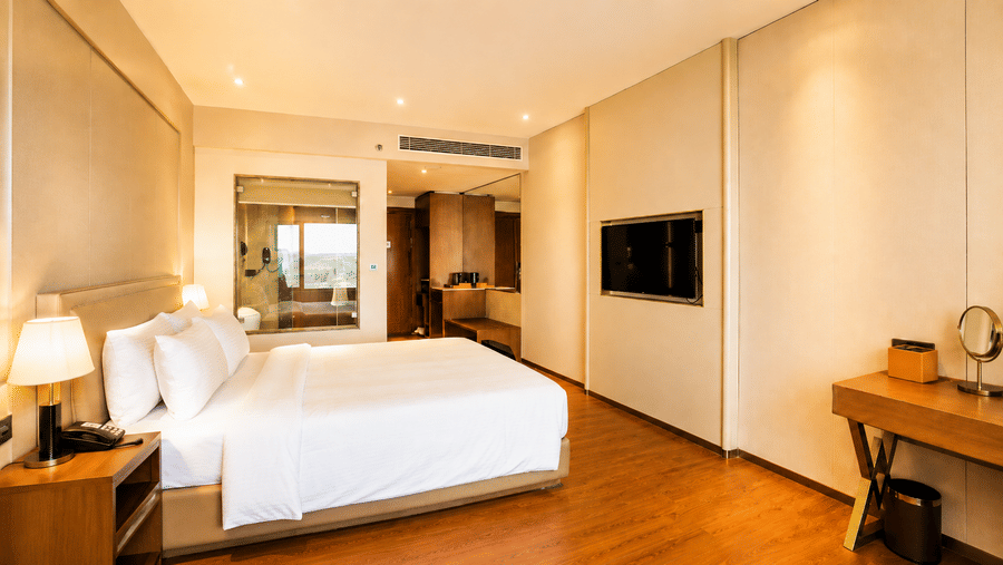 Superior King Room at Pride Plaza Indore showing a king bed with pillows, a bedside table with lamp, a wall-mounted television, and a desk area.