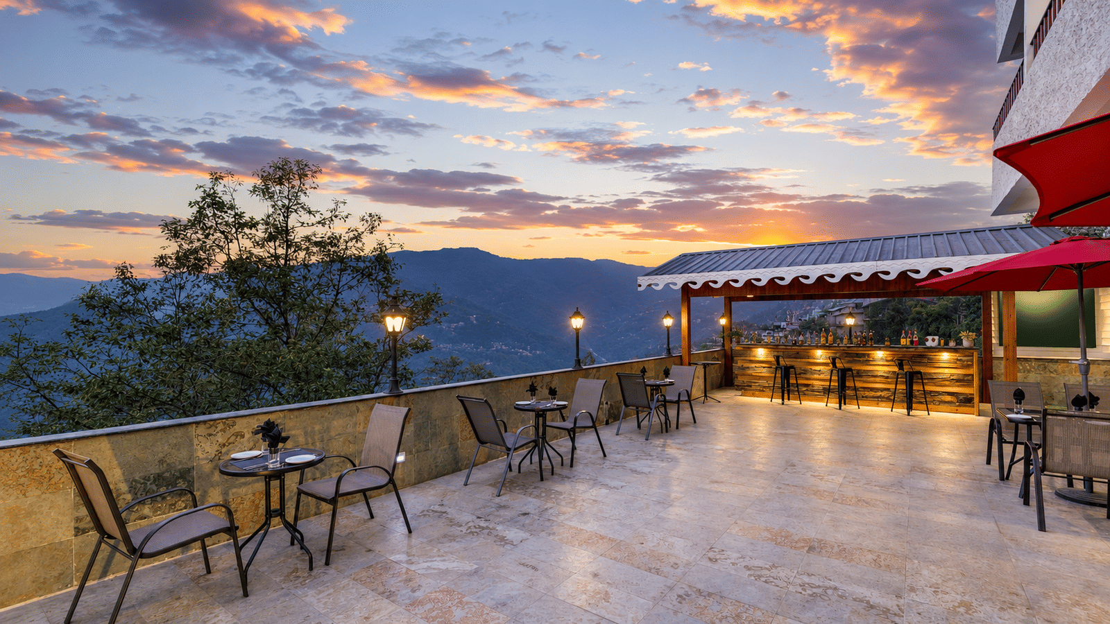 Terrace bar at Valmere Himalayan Hotel & Spa, Gangtok at sunset, with 3 wooden barstools, patio seating, red umbrellas, and mountain views beyond the railing.