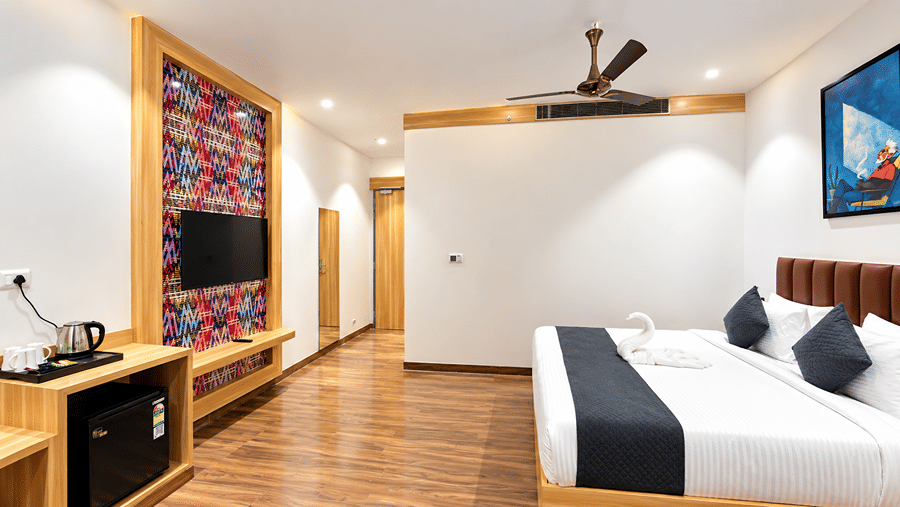 Room at Daksh The Royal Braj, Vrindavan, with a bed, a wall-mounted television on a colourful panel, a black cabinet, and a ceiling fan.
