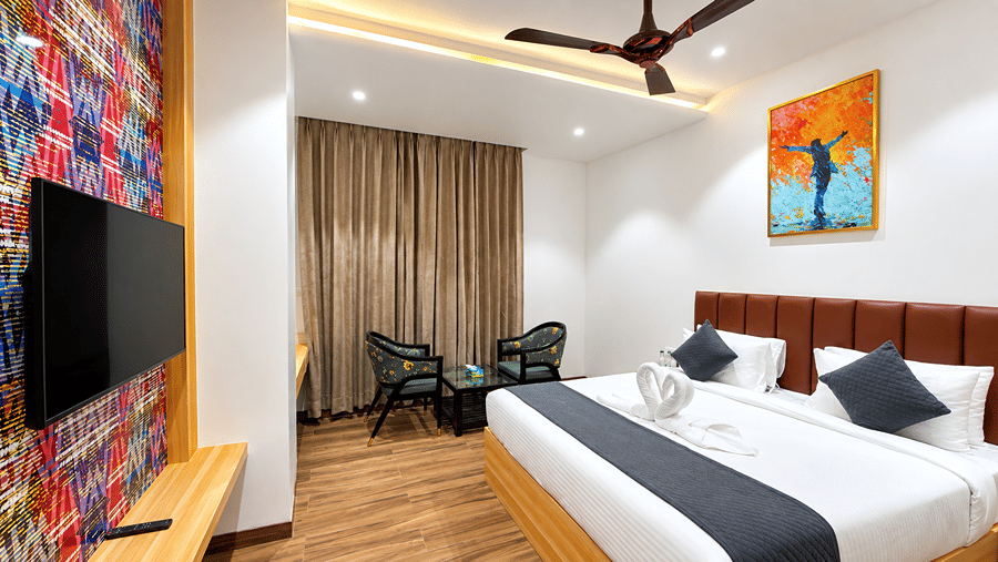 Room at Daksh The Royal Braj, Vrindavan, with a bed, a wall-mounted television, two armchairs, curtains, and a ceiling fan.
