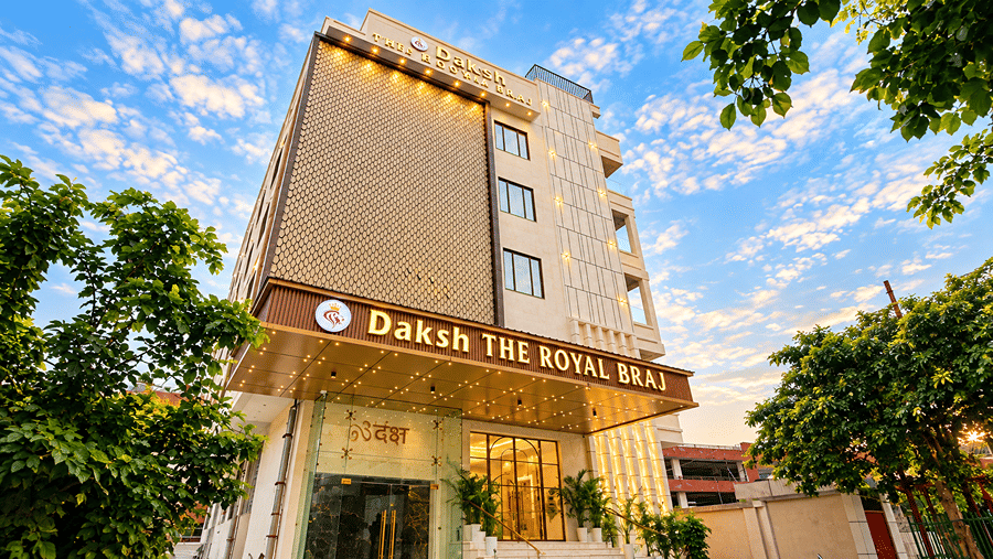 Angled view of the multi-storey facade of Daksh The Royal Braj, Vrindavan, with hotel signage and trees beside the building.
