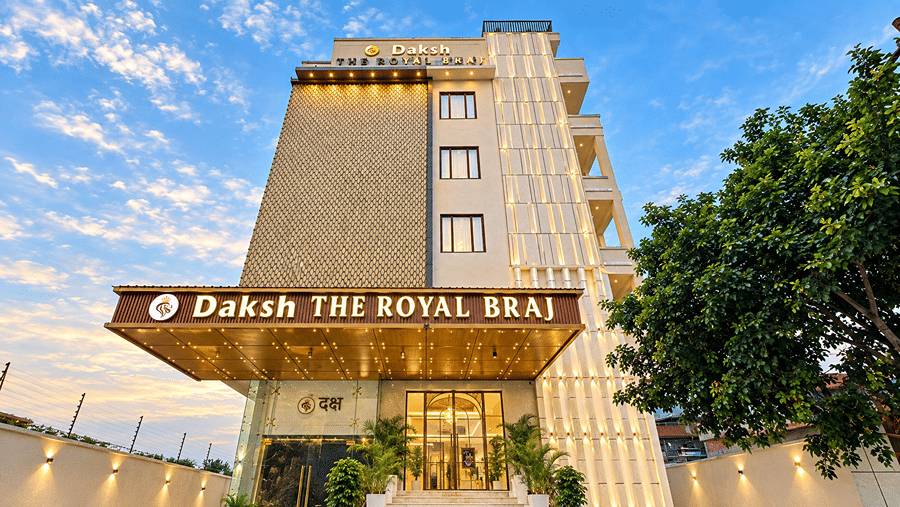Front facade of Daksh The Royal Braj, Vrindavan, showing a multi-storey building with hotel signage, an entrance canopy, and trees under a blue sky.