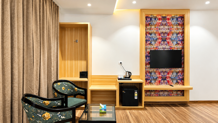 Room at Daksh The Royal Braj, Vrindavan, with a wooden desk, a wall-mounted television on a colourful panel, an armchair, and curtains.