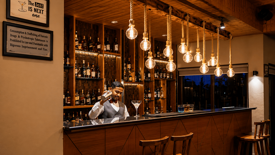 A bartender at Hotel Sonar Bangla, Sundarban mixing a drink behind a wooden bar counter with hanging lights. 