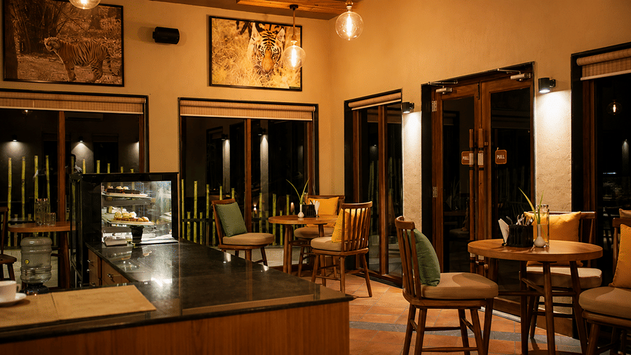 A cafe seating area at Hotel Sonar Bangla, Sundarban featuring wooden tables, chairs with cushions, and a display counter. 