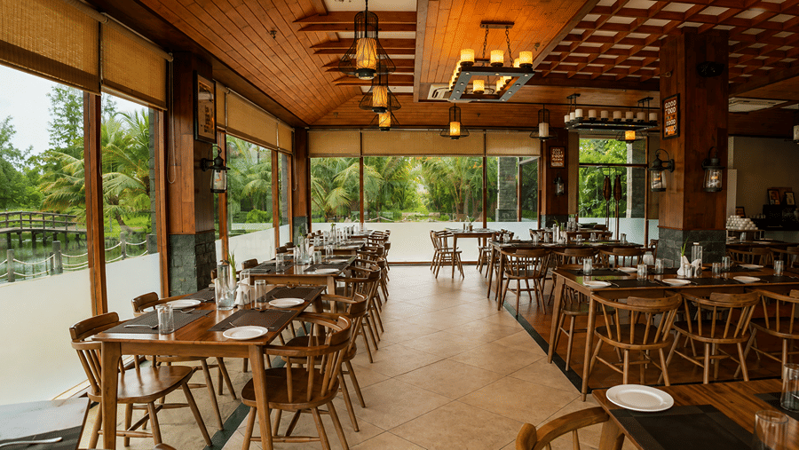 An interior view of a dining hall at Hotel Sonar Bangla, Sundarban featuring wooden tables, chairs, and hanging lamps. 