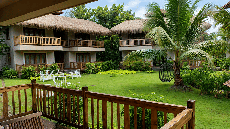 An exterior view of cottages at Hotel Sonar Bangla, Sundarban featuring thatched roofs, wooden balconies, lawn, and outdoor seating. 
