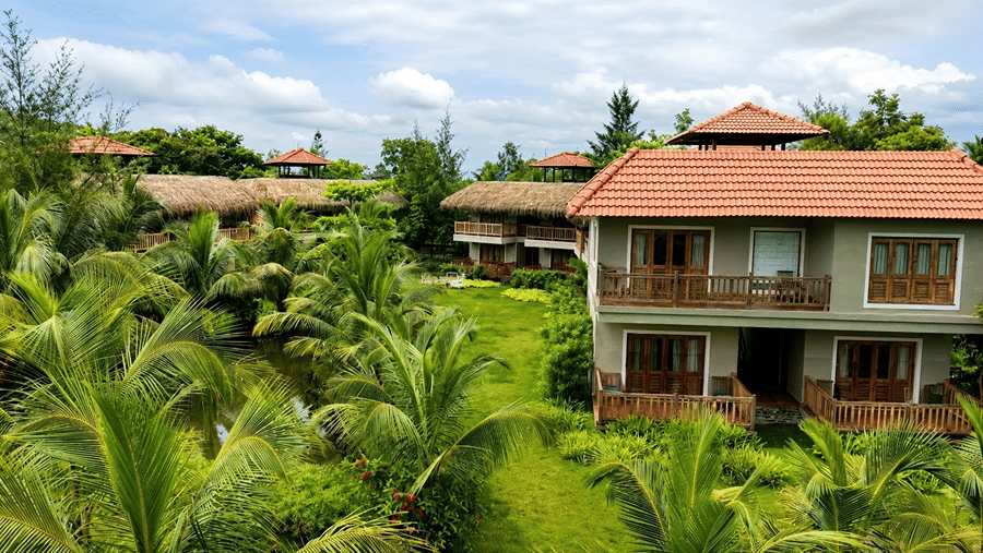A view of cottages at Hotel Sonar Bangla, Sundarban featuring tiled roofs, palm trees, pathways, and lawns. 