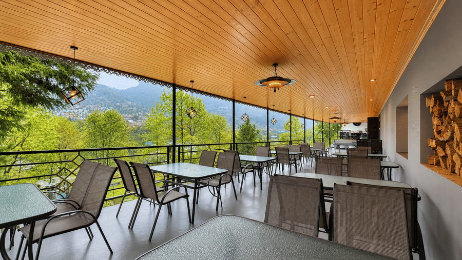 Opal bar at Orsino Khangri Karpo Retreat & Spa, Lachung featuring rectangular tables and chairs are arranged along a balcony. The view shows a valley with trees and mountain peaks.
