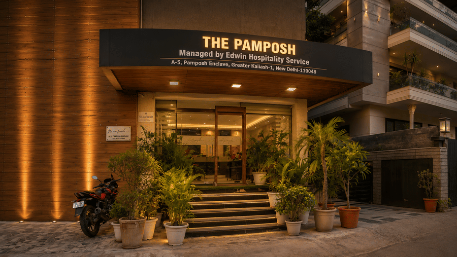 The illuminated facade of The Pamposh, New Delhi, with plants and the sign board.
