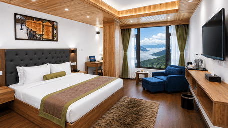 Guest room at Valmere Magnolia Boutique Hotel & Spa, Pelling with a bed, dark headboard, a navy armchair and ottoman, and a window overlooking clouds and hills.