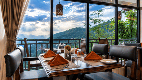 Restaurant table at Valmere Magnolia Boutique Hotel & Spa, Pelling set with orange napkins and glassware, beside windows overlooking green mountains.