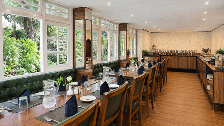 Restaurant at Valmere Thistle Boutique Hotel, Darjeeling with dark wood tables, green tufted banquette seating, and large windows facing greenery.