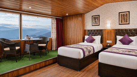 Twin beds at Valmere Thistle Boutique Hotel, Darjeeling beside a window seating nook with wicker chairs overlooking layered mountain ranges.