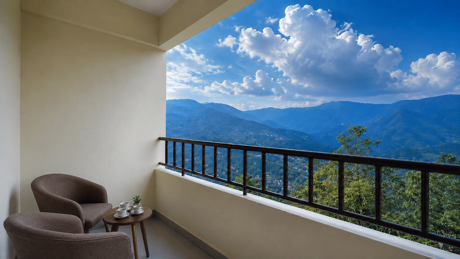 Balcony at Valmere Himalayan Hotel & Spa, Gangtok with 2 armchairs, a round table with cups, and a sweeping view of forested mountains under a partly cloudy sky.