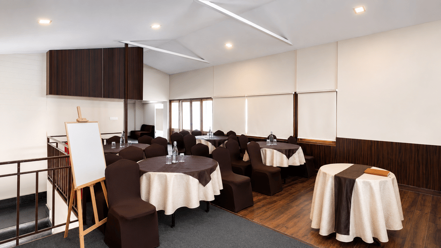 Conference room at Valmere Himalayan Hotel & Spa, Gangtok set up with round tables, brown chair covers, water bottles, and a whiteboard resting on an easel.