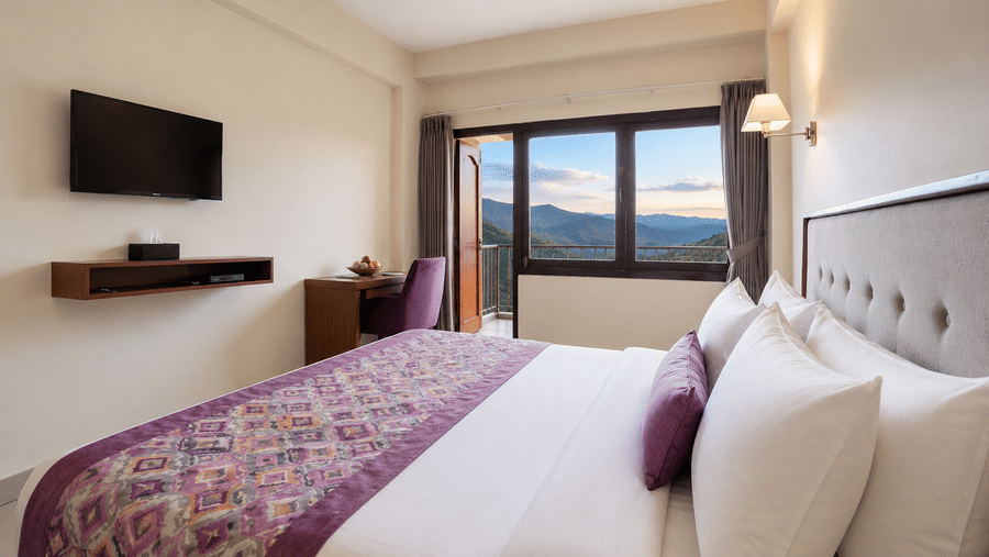 Deluxe Room at Valmere Himalayan Hotel & Spa, Gangtok with a king-size bed, purple bolster pillow, and an open balcony door revealing a mountain sunrise view.