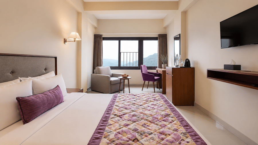 Deluxe Room at Valmere Himalayan Hotel & Spa, Gangtok with a king-size bed, purple pillow, a grey armchair and purple chair by the window, and a wall-mounted TV.