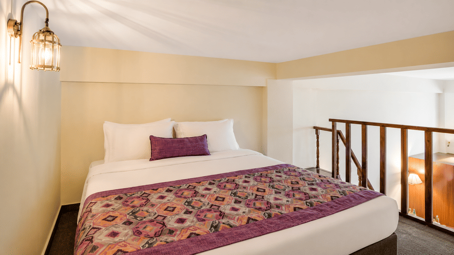 Mezzanine loft bed at Valmere Himalayan Hotel & Spa, Gangtok with white bedding, a purple bolster pillow, a wooden railing, and a view down to the room below.