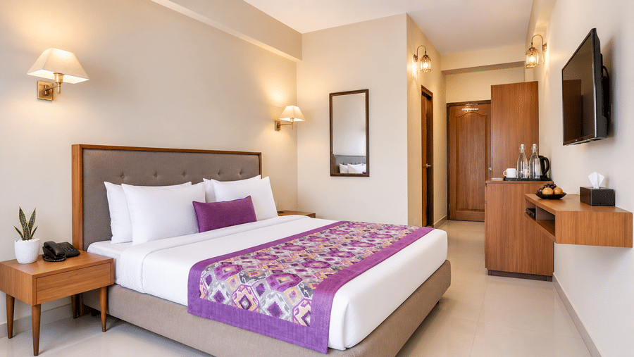 Guest room at Valmere Himalayan Hotel & Spa, Gangtok with a king-size bed, purple bolster pillow, a wall mirror, and a hallway leading to a wooden dresser and TV.
