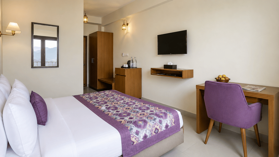 Guest room at Valmere Himalayan Hotel & Spa, Gangtok with a king-size bed, a wall mirror reflecting mountain views, and a desk with a purple chair and newspaper.