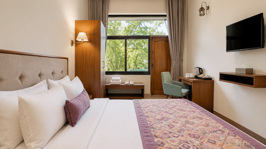 Guest room at Valmere Himalayan Hotel & Spa, Gangtok with a king-size bed, a wooden wardrobe, a teal armchair at a work desk, and a window overlooking trees.
