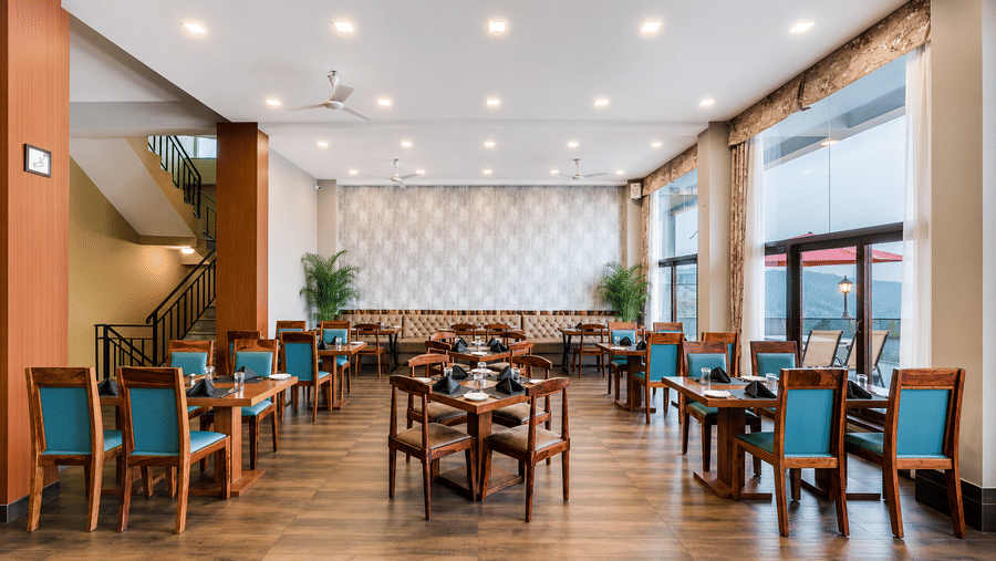 Restaurant at Valmere Himalayan Hotel & Spa, Gangtok with wooden chairs upholstered in teal, set tables, a beige banquette, and large windows to a terrace.