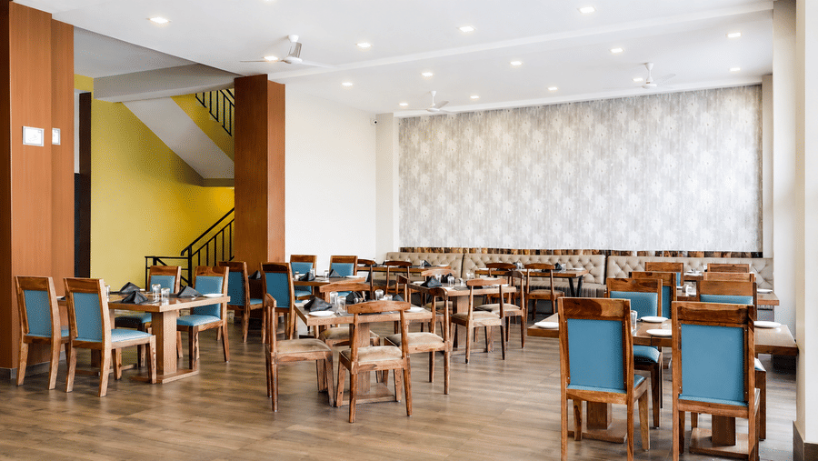 Restaurant at Valmere Himalayan Hotel & Spa, Gangtok with wooden chairs in teal upholstery, set tables, a yellow accent wall, and a staircase in the background.