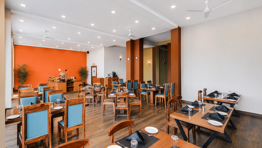 Restaurant at Valmere Himalayan Hotel & Spa, Gangtok with wooden tables set with plates and napkins, teal chairs, and a buffet counter against an orange wall.
